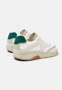 Paire de baskets blanches avec des accents beige et vert, semelles épaisses beiges et détails jaunes sur la bande de roulement, vues de dos et de profil.
