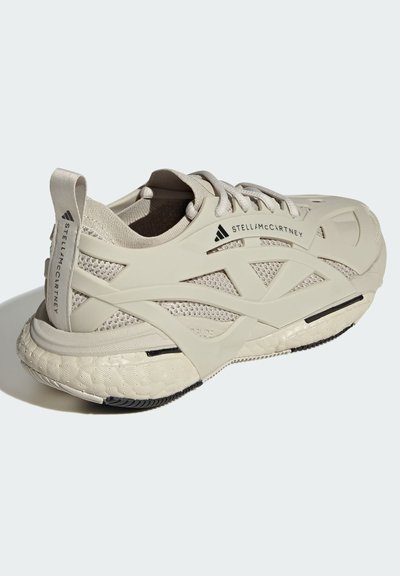 Zapato deportivo de color beige claro con una parte superior de malla texturizada, suela de goma y un dibujo pronunciado. Presenta un diseño minimalista y acentos de marca.