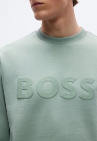 Világoszöld pulóver bordázott környakú résszel és azonos színű, kiemelkedő "BOSS" logóval az elülső részen. Sima felületű anyag.