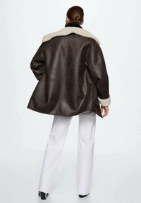 Giacca di pelle marrone con un colletto in pelliccia sintetica color crema, vestibilità morbida e cuciture posteriori visibili. Abbinata a pantaloni bianchi, che mostrano una silhouette rilassata.