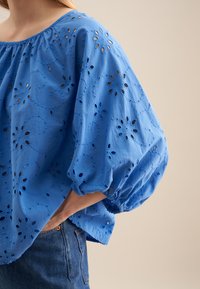 Blå bomuldsbluse med puffede ærmer, der har blomsterbroderi og udskæringsmønstre. Sammen med denimjeans.