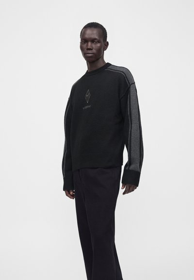 Homme portant un pull noir avec des rayures grises sur les manches et un logo géométrique sur la poitrine, associé à un pantalon noir, debout sur un fond blanc.