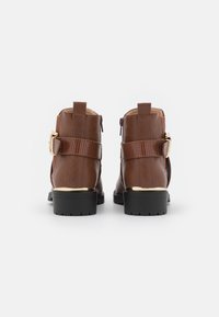 Hnědé kožené kotníkové boty s texturovaným páskem, zlatými akcenty a bočními zipy. Pevná černá gumová podrážka s protiskluzovými drážkami.