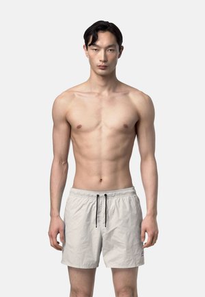 Joven con cabello corto y oscuro que lleva puestos unos shorts de baño beige claros con cordones negros, de pie mirando de frente sobre un fondo liso.