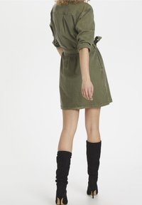 Olivově zelené šaty s páskem a ohrnutými rukávy, nad kolena, se sklady na zádech. V kombinaci s černými semišovými kozačkami po kolena.