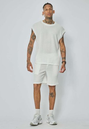 Homme avec des tatouages portant un haut blanc texturé sans manches et un short assorti avec des baskets blanches, debout devant un fond clair uni.