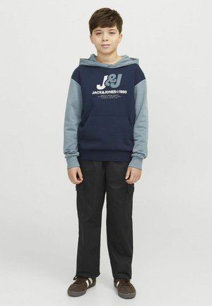 Granatowy hoodie z szarymi rękawami, z białym logo. W połączeniu z czarnymi spodniami cargo i brązowymi butami z białymi paskami.