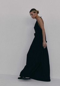 Czarna, bezrękawnikowa sukienka maxi o luźnym kroju, z delikatną teksturą i odkrytymi plecami. Noszona z czarnymi płaskimi butami.