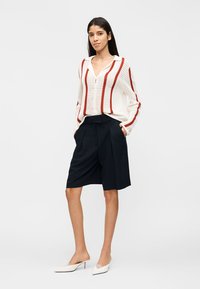 Γυναίκα που φοράει λευκό πλεκτό πουλόβερ με κόκκινες ρίγες, μαύρο σορτς μέχρι το γόνατο και λευκά παπούτσια με χαμηλό τακούνι, στέκεται με τα χέρια στις τσέπες.