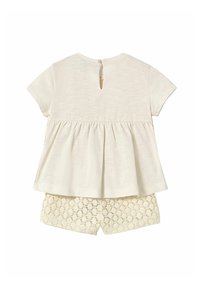 Top peplum in tessuto crema con maniche corte e chiusura posteriore con bottoni, abbinato a pantaloni corti in pizzo bianco con orlo a scallop.