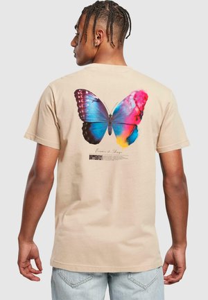 T-shirt en coton couleur crème avec un grand motif de papillon vibrant en bleu et rose au dos, accompagné de texte en dessous du design.