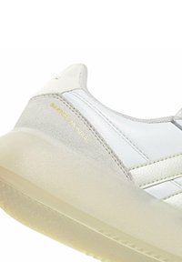 Gros plan sur une basket blanche avec un talon en daim beige, une semelle épaisse translucide et un texte doré "Barreda Mundial" sur le côté.