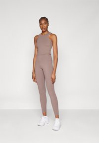 Bruin activewear set met een fitted mouwloos shirt en high-waisted leggings. De stof is soepel met een lichte glans. Witte sportschoenen maken de look compleet.