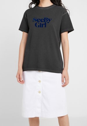 Kobieta w czarnym t-shircie z napisem „See By Girl” i białej spódnicy z guzikiem z przodu, stojąca na tle jasnoszarego tła.