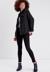 Musta teksturoitu takki, jossa on kaulus ja etunapit, yhdistetty mustiin istuviin farkkuihin ja nilkkureihin. Malli seisoo neutraalissa taustassa.