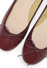 Ballerine bordeaux con punta tonda, dettaglio a fiocco sottile e texture in pelle liscia. L'interno presenta una fodera beige con una leggera nervatura.