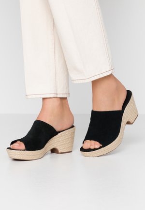 Fødder iført sorte ruskinds kilehælssandaler med åbne tæer og flettede platformssåler, kombineret med off-white korte bukser mod en ensfarvet baggrund.