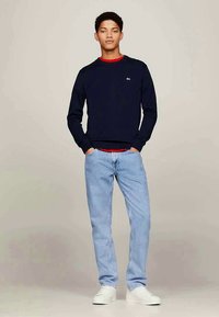 Giovane in piedi, mani nelle tasche, indossa un maglione blu navy su una camicia rossa, jeans azzurri chiari e sneakers bianche su uno sfondo neutro.