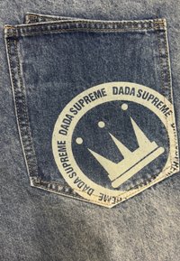 Un bolsillo trasero de tela vaquera que presenta un gran parche redondo color crema con el texto "DADA SUPREME" y un diseño de corona, contorneado en azul oscuro.