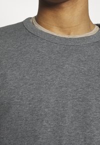 Detailní záběr na krk a horní část hrudníku osoby v šedém tričku s kulatým výstřihem, s viditelným límcem béžového pruhovaného trička.