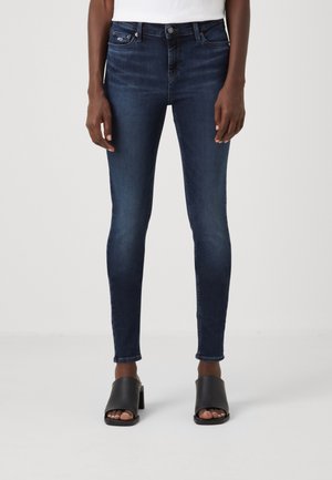 Ciemnoniebieskie obcisłe dżinsy noszone przez stojącą osobę, zestawione z czarnymi sandałami na obcasie z odkrytymi palcami, na jednolitym jasnym tle.