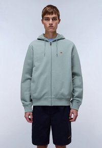 Hele-roheline lukuga dressipluus esi lukuga ja kapuutsiga, millel on väike logo rinnal, kombineeritud tumesiniste lühikestega.