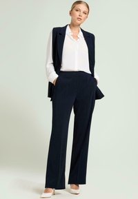 Marineblauwe op maat gemaakte broek met een bijpassend mouwloos colbert, gedragen over een witte overhemd met knopen. De broek heeft rechte pijpen en zakken. Geaccentueerd met witte hakken.