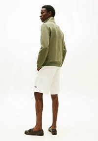 Homme debout les mains dans les poches, portant une veste zippée vert clair, un short blanc et des mocassins marron foncé, sur un fond blanc uni.