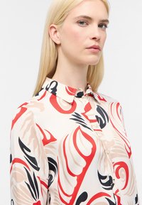 Blusa fluida in tessuto crema con un audace motivo astratto in rosso, nero e beige. Collo classico con bottoni sul davanti.