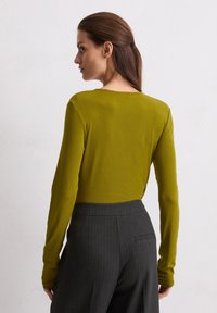 Olivengrünes Langarmoberteil aus glattem Stoff mit rundem Ausschnitt. Kombiniert mit dunkelgrauen, gestreiften High-Waist-Hosen.