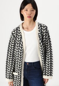 Cardigan em preto e creme com padrão de espinha de peixe, apresentando uma textura de malha grossa, frente aberta e colarinho canelado.