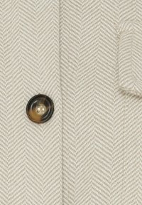 Beige hørfisket stof med en synlig sort og ravfarvet knap. Har et tekstureret vævemønster og en sidelomme detalje.