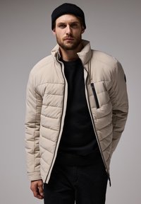 Beige isolierte Jacke mit gestepptem Muster, hohem Kragen, Reißverschlusstasche und schwarzen Akzenten, getragen über einem schwarzen Pullover.