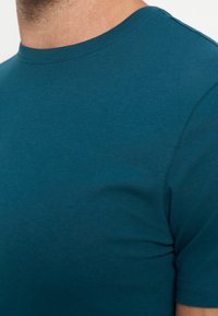 Close-up af en person iført en blågrøn t-shirt med korte ærmer og rund hals, der viser den øverste bryst- og halsområde.