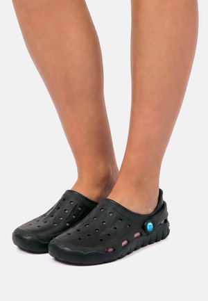 Persona que lleva puestos zuecos negros sin cordones con agujeros de ventilación y un logo circular azul en el lateral, sobre un fondo claro.