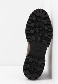 Semelle extérieure en caoutchouc noir avec des crampons profonds pour l'adhérence, surface texturée et design robuste, fixée à un dessus en daim marron.