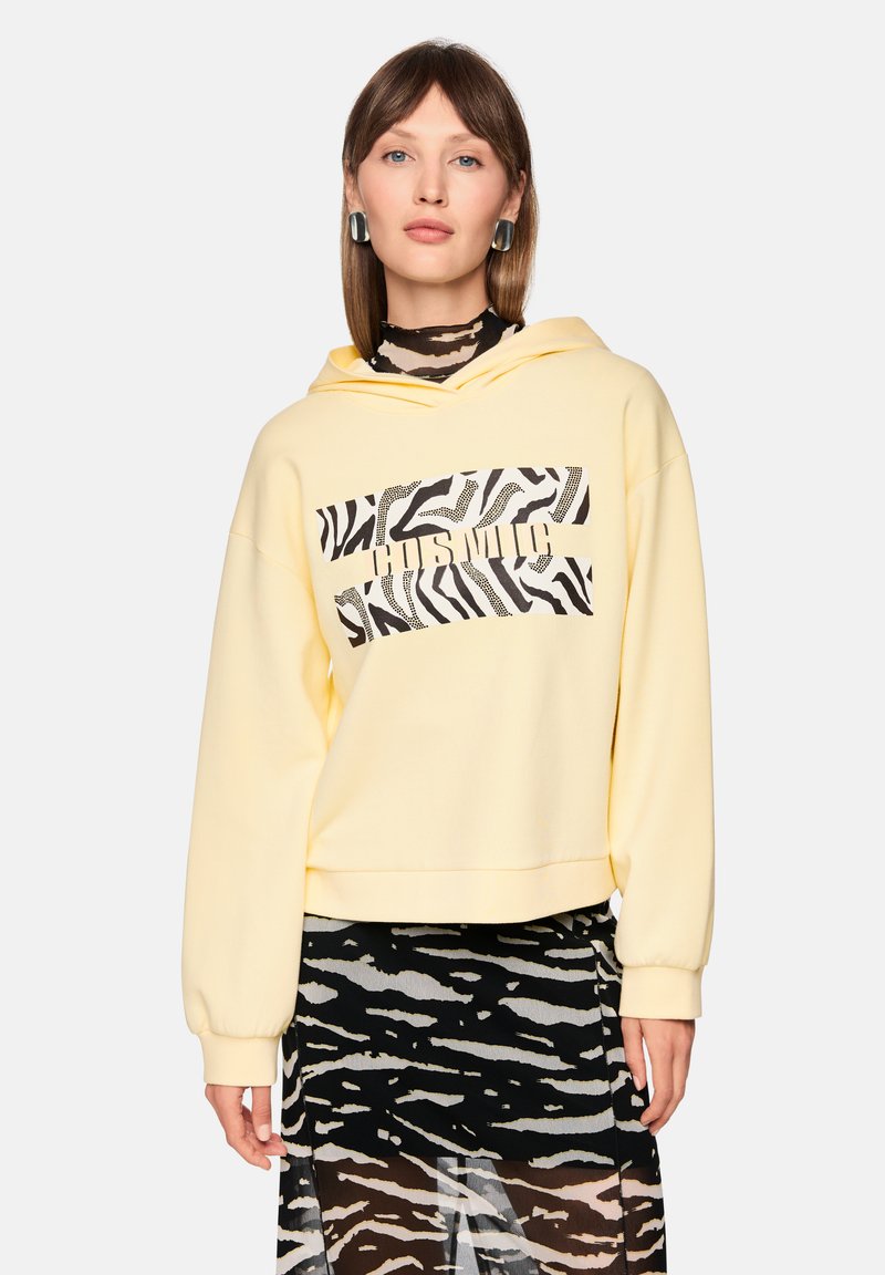 Gelber kurzer Hoodie mit Kapuze, die ein schwarz-weißes Zebra-Muster aufweist. Der Stoff wirkt weich und hat gerippte Bündchen.