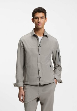 Hellgraue Jacke zum Knöpfen aus strukturiertem Stoff, mit langen Ärmeln und einer Taschenapplikation am linken Arm. Kombiniert mit passenden hellgrauen Hosen.