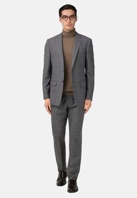 Chaqueta de traje de cuadros grises con pantalones a juego, llevada sobre un suéter de cuello alto marrón. Zapatos marrones completan el conjunto. Líneas limpias y diseño ajustado.