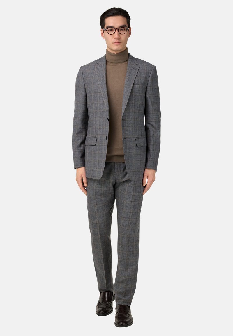 Chaqueta de traje de cuadros grises con pantalones a juego, llevada sobre un suéter de cuello alto marrón. Zapatos marrones completan el conjunto. Líneas limpias y diseño ajustado.