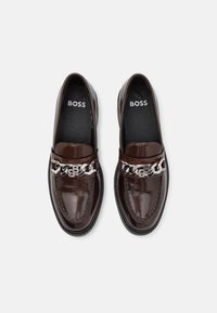 Mocassins en cuir marron avec une finition brillante, un accent de chaîne sur le dessus et des semelles intérieures noires imprimées du nom de la marque.