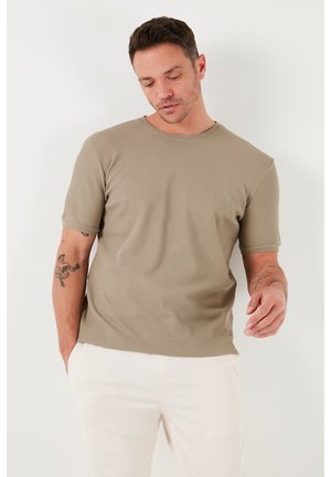 Beige, rövid ujjú póló, textúrázott bordás anyagból, kerek nyakkivágással, testhezálló sziluettel, világos színű nadrággal viselve; látható kar tetoválással.