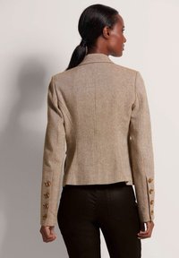 Beige op maat gemaakte blazer van een gestructureerde stof, met een enkele knoopsluiting en goudkleurige knopen op de manchetten. Getailleerde silhouet, achteraanzicht.