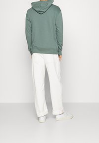 Felpa verde con cappuccio e tasca posteriore, vestibilità comoda. Abbinata a pantaloni bianchi larghi e sneakers chiare. Tessuto liscio, design semplice.