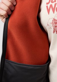 Fleece-Weste mit einem strukturierten, leuchtend orangefarbenen Innenfutter und einem glatten, dunkelgrauen Außenmaterial, ausgestattet mit einem schwarzen Reißverschluss und kontrastierendem Taschenrand.