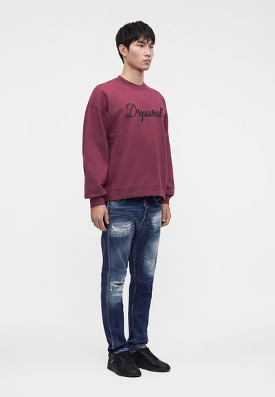 Νεαρός άνδρας με μπορντό φούτερ Dsquared2, παλιωμένο μπλε τζιν και μαύρα αθλητικά παπούτσια, στέκεται μπροστά σε απλό λευκό φόντο.