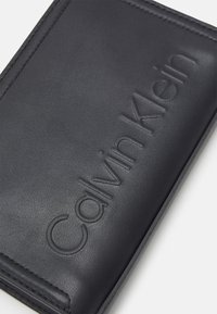 Portafoglio in pelle nera con logo "Calvin Klein" in rilievo. Texture liscia, forma rettangolare e bordi cuciti per una maggiore durata.
