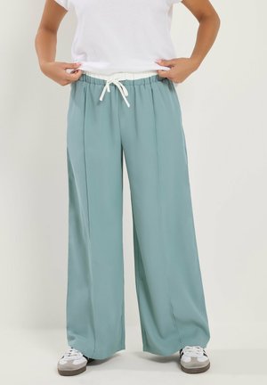 Persona usando pantalones anchos de pierna en azul claro con cordón blanco y zapatillas blancas, ajustándose la cintura del pantalón.