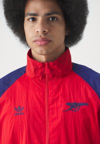 Crvena i mornarsko plava jakna na zip, lagani materijal, s izvezenim logotipima na prsima, visokim ovratnikom i teksturiranom površinom.