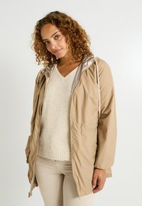 Femme aux cheveux bouclés portant une veste à capuche beige sur un pull crème et un pantalon beige clair, se tenant devant un fond blanc.